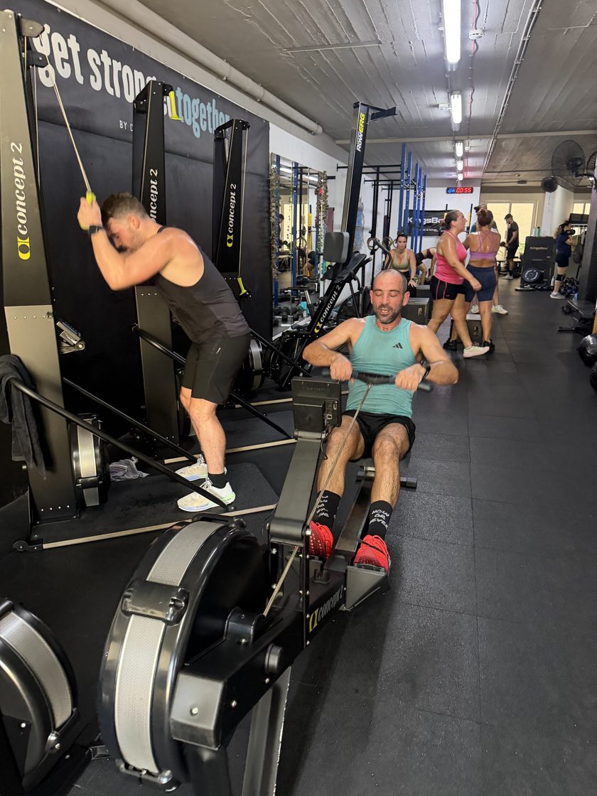 Two men training side by side, one on a ski erg and one on a rower, with members working out on the floor behind them